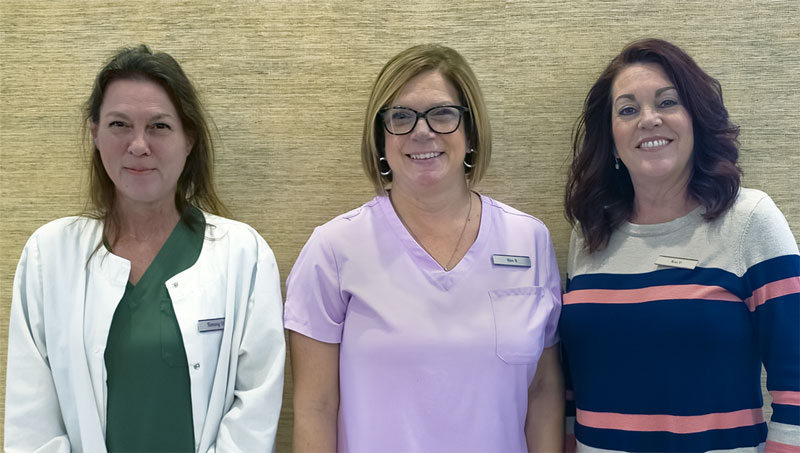 Three staff members of Peaceful Harbor Gynecology with standing side by side, smiling at the camera. The woman on the left is wearing a white coat, the middle woman is in a light purple top, and the woman on the right is in a black and pink striped sweater. They are in front of a beige background.